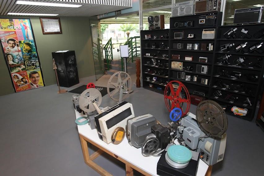 área interna de museu. com mesa e vários objetos antigos, como câmeras e filmadoras sobre uma mesa. Ao fundo, prateleiras com objetos de fotografia. #paratodosverem