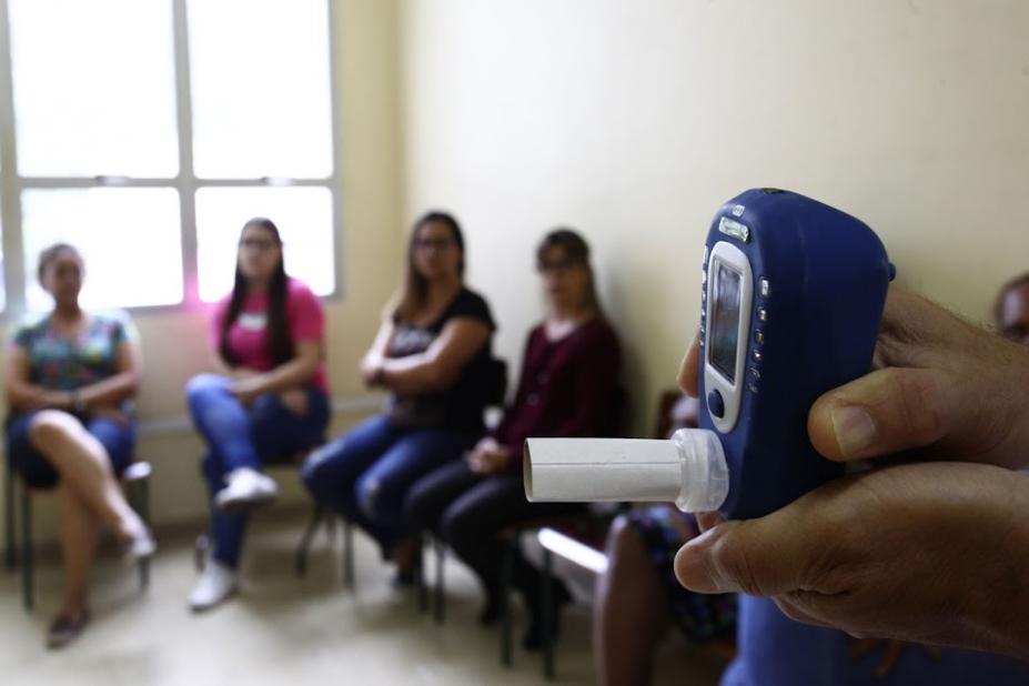 monoxímetro nas mãos de uma pessoa em primeiro plano à direita. Ao fundo, desfocadas, há mulheres sentadas em cadeiras. #paratodosverem