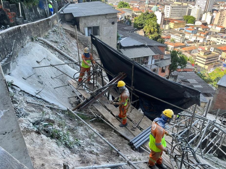 homens estão trabahando em encosta em meio a estruturas de ferro. Há moradias abaixo. #paratodosverem