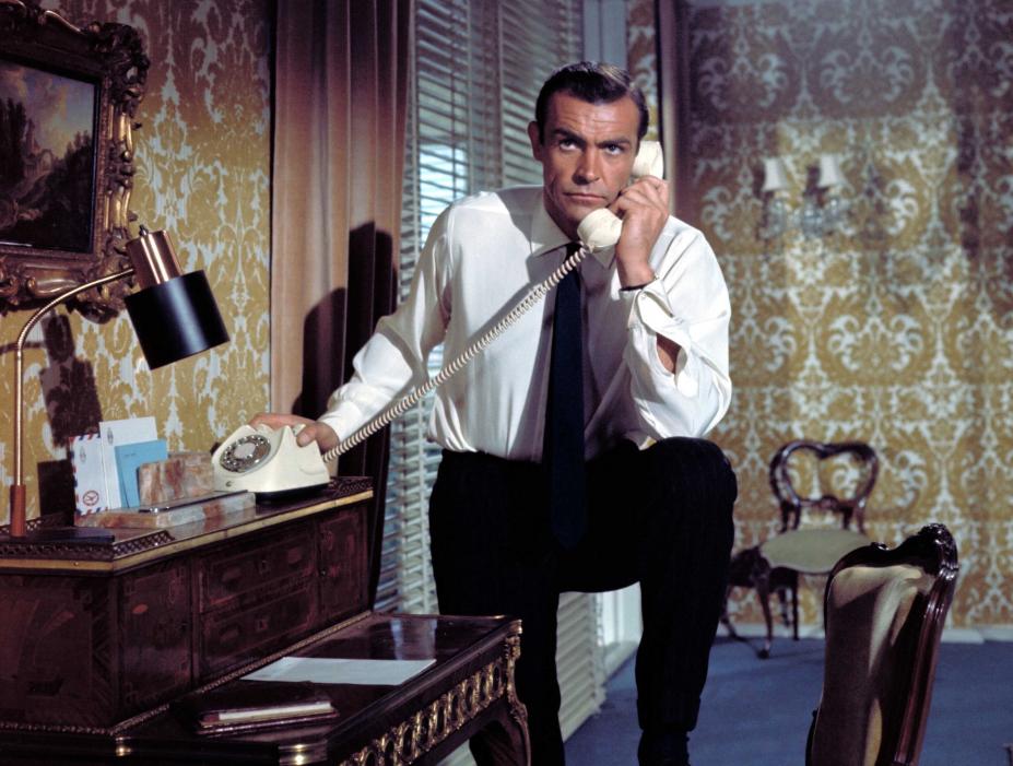 James Bonde está ao telefone em um quarto. Uma de suas pernas está apoiada sobre uma cadeira. #Pracegover