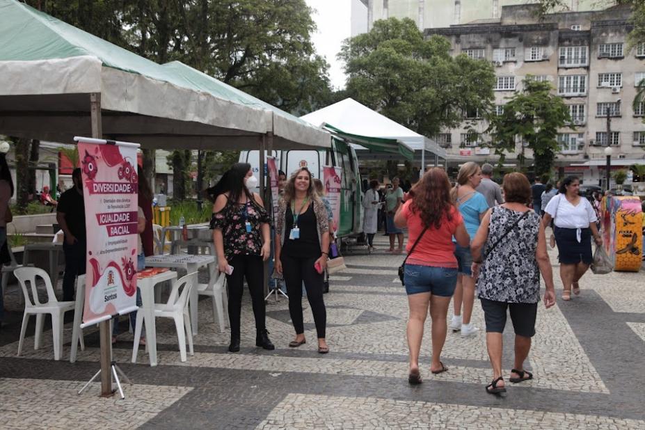 tendas armadas na praça à esquerda. Mulheres em frente às tendas e circulando. #paratodosverem
