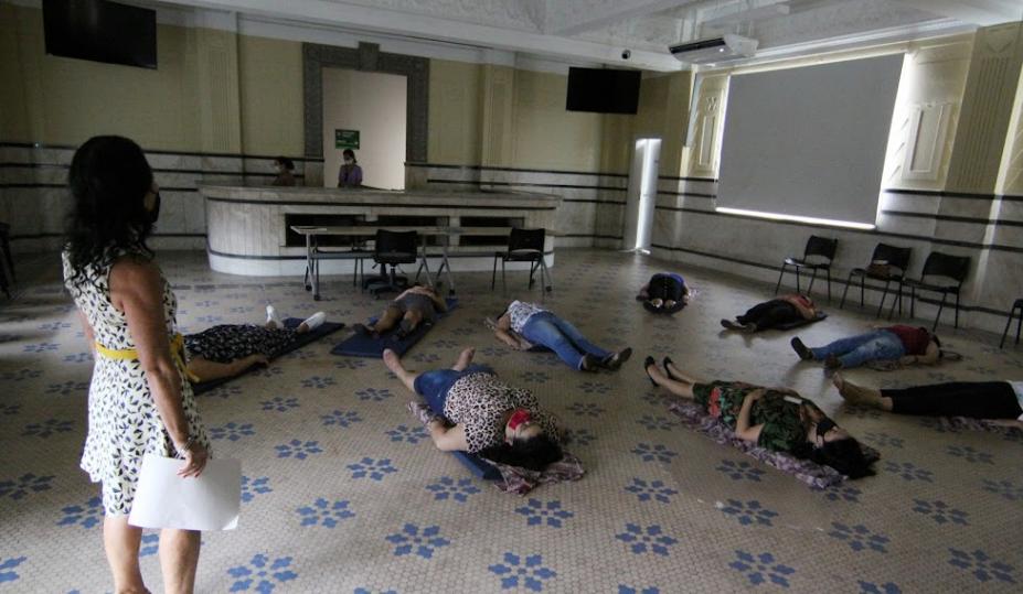 mulheres estão deitadas no chão sobre colchonetes. Uma está em pé como coordenadora da atividade. O ambiente é fechado a luz está apagada. #paratodosverem