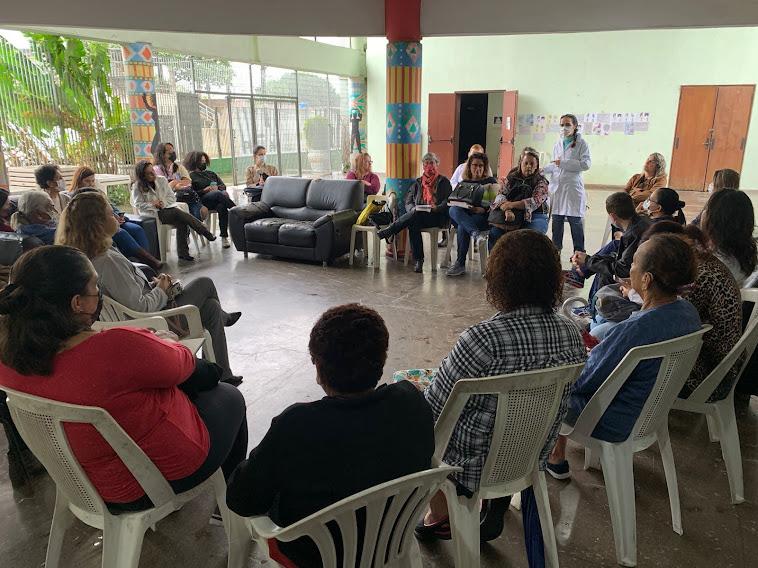 mulheres estão sentada em cadeiras, em formação de roda. Uma delas, ao fundo, está em pé falando a todas. #paratodosverem