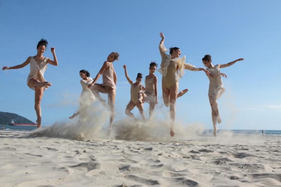 Bailarinas saltam na areia em dia ensolarado sem nuvens no céu azul ao fundo. #pracegover