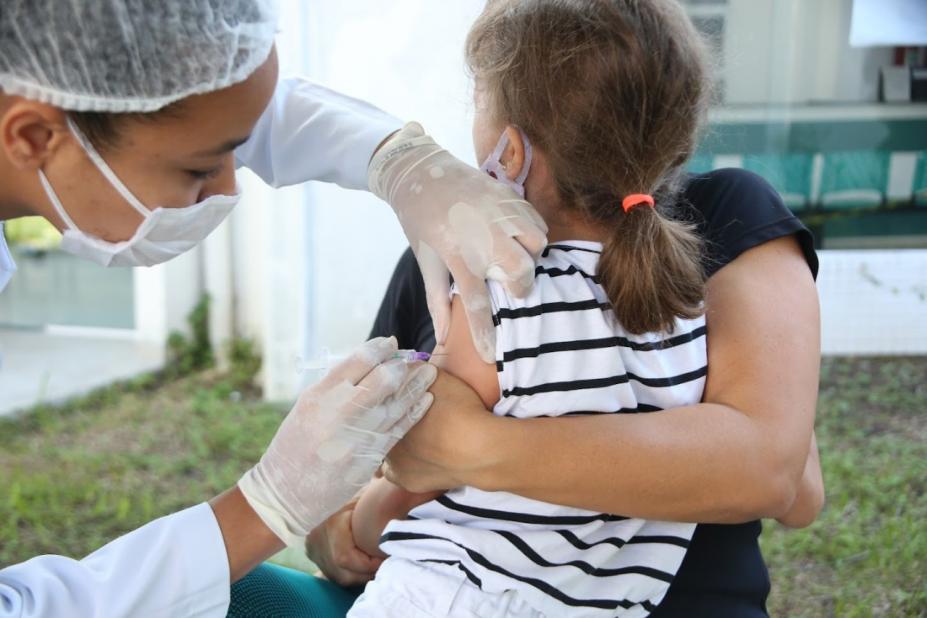 menina, de costas para a foto, está no colo da mãe sendo vacinada por uma mulher. #paratodosverem