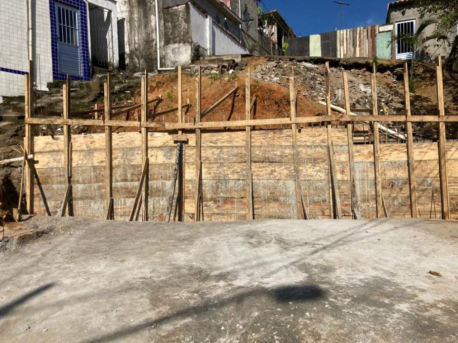 estrutura em madeira armada para construção de muro. Há um trecho em aclive acima e casas ao lado esquerdo e ao fundo. #paratodosverem