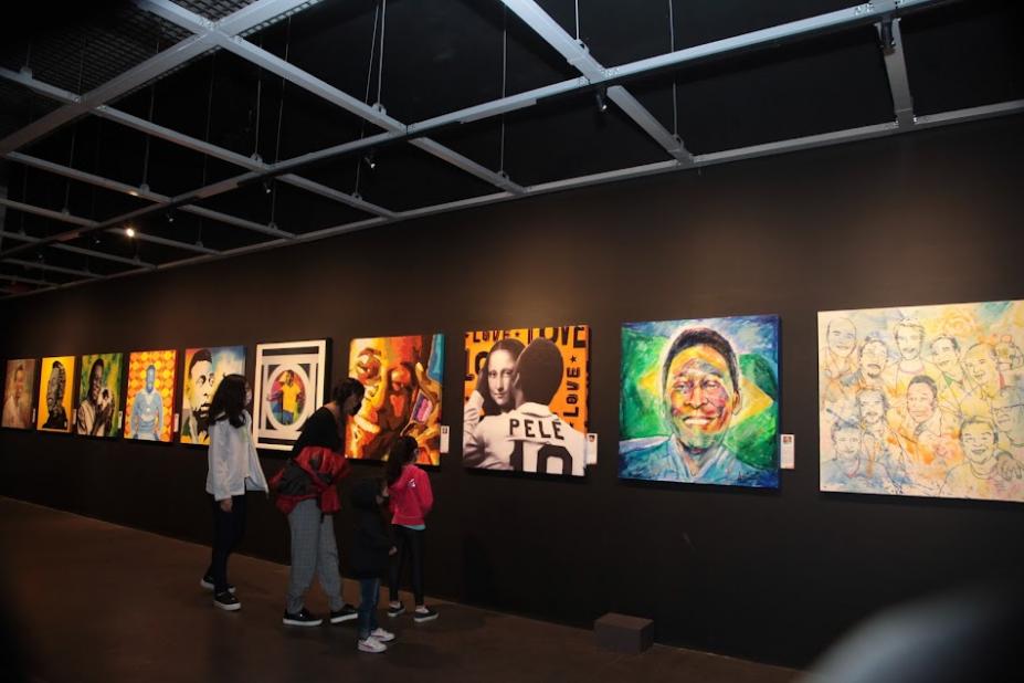 Interior do Museu Pelé com fotos e pinturas do Rei feitas por artistas. Há público observando. #paratodosverem