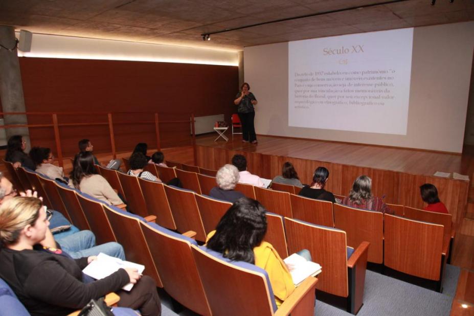 Mulher dá palestra sobre palco de auditório do Museu Pelé, assistido por público sentado. #pracegover