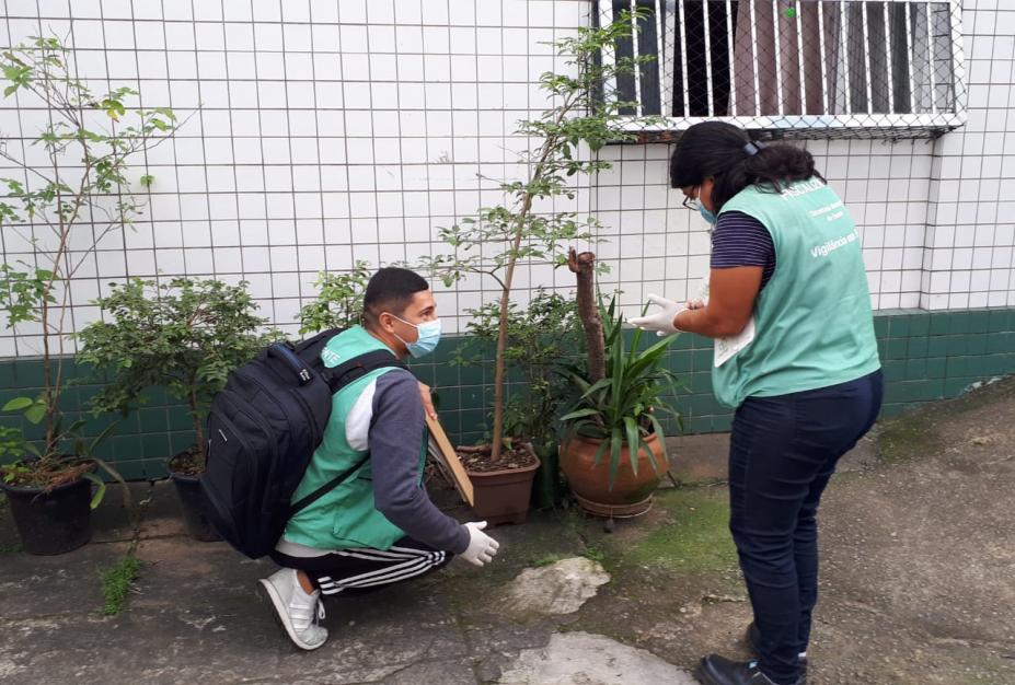 dois agentes estão no quintal de uma moradia. O rapaz está agachado próxima a alguns vasos. A moça está em pé fazendo anotação em uma prancheta. #paratodosverem