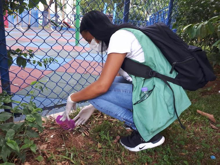 Mulher usando luvas e jaleco esta ajoelhada em gramado mexendo na terra. #Paratodosverem