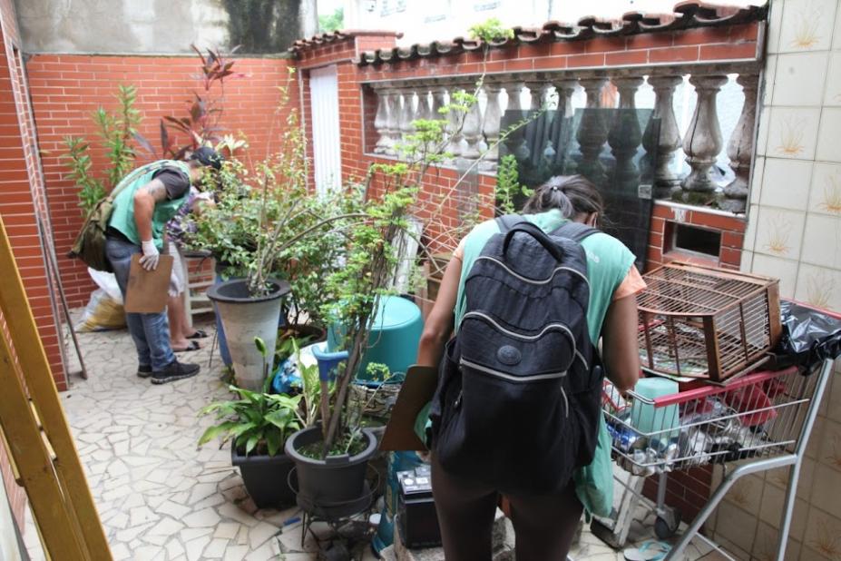 Agentes estão na parte externa da frente de uma casa onde procuram focos de larvas. A área tem muitas plantas e quinquilharias. #paratodosverem