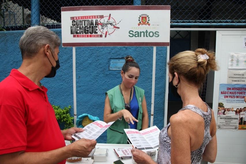 duas pessoas estão segurando folders informativos diante de estande. Estande tem placa onde se lê Todos contra a dengue. Uma mulher usando um colete de identificação está falando com as duas pessoas. #paratodosverem