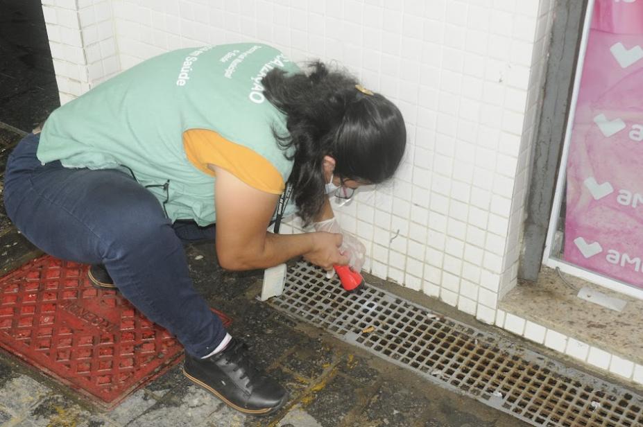 agente uniformizada com colete de identificação aponta lanterna para grelha no piso. #paratodosverem