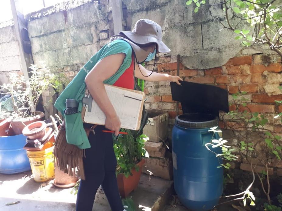 Agente uniformizada mexe em galão encostado em muro. Vários vasos estão junto à parede. #Paratodosverem