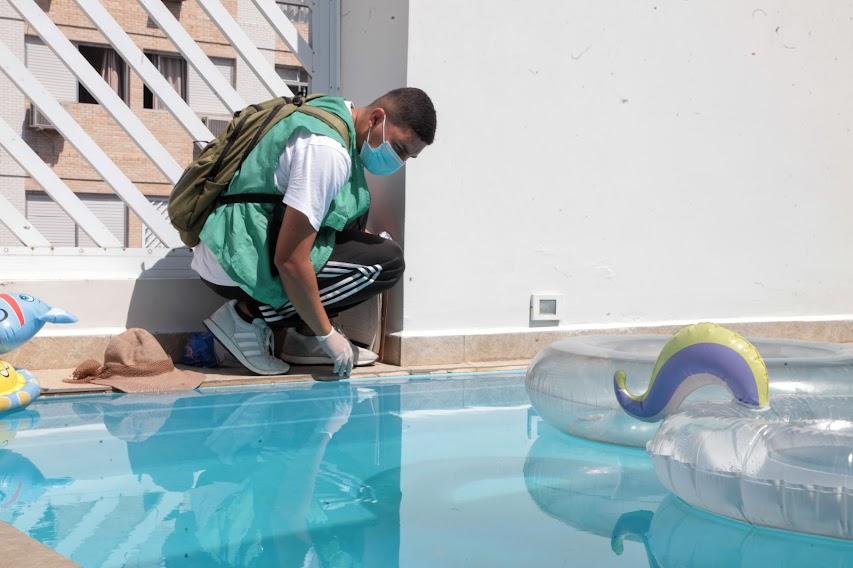 agente uniformizado se ajoelha ao lado de piscina e aproxima frasco da linha d'água. Há boias ao lado direito da piscina. #paratodosverem