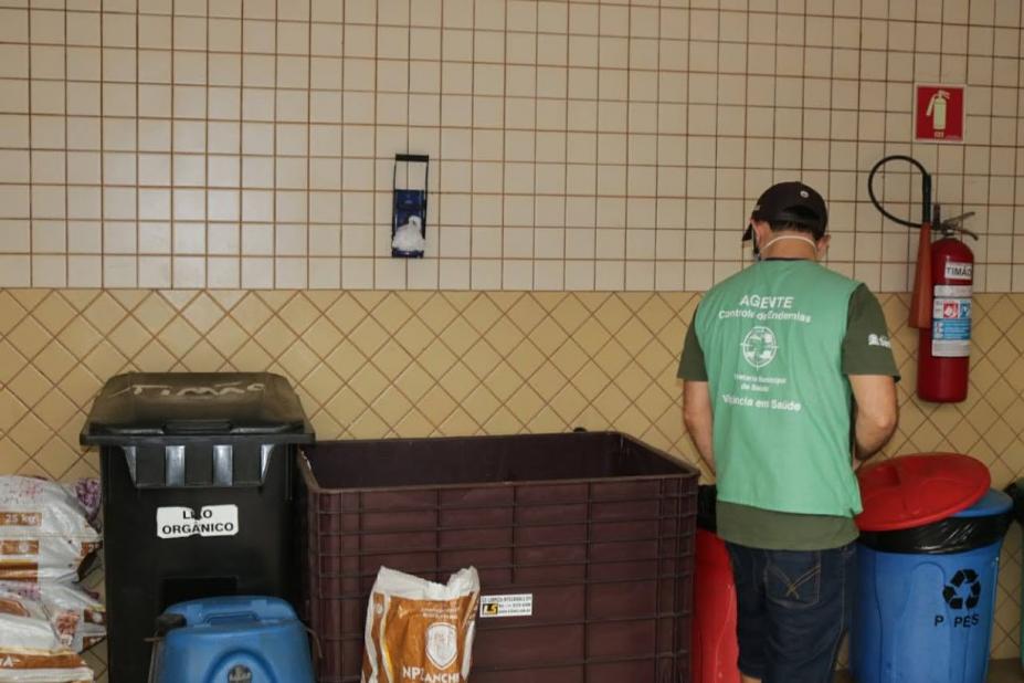 Agente esta ao lado de contêineres de lixo orgânico e reciclável. #Paratodosverem