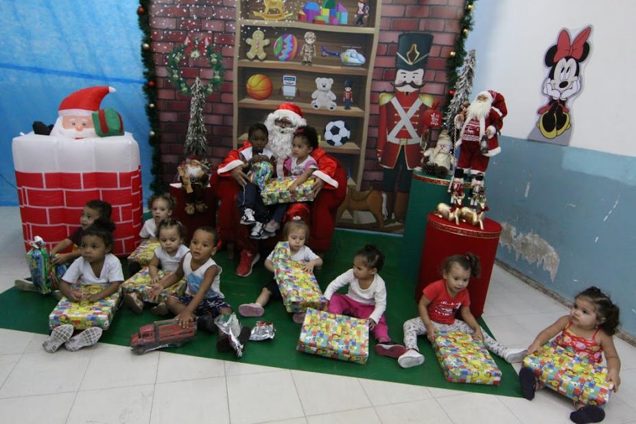 crianças segurando brinquedos e papai noel ao fundo com duas crianças no colo #paratodosverem