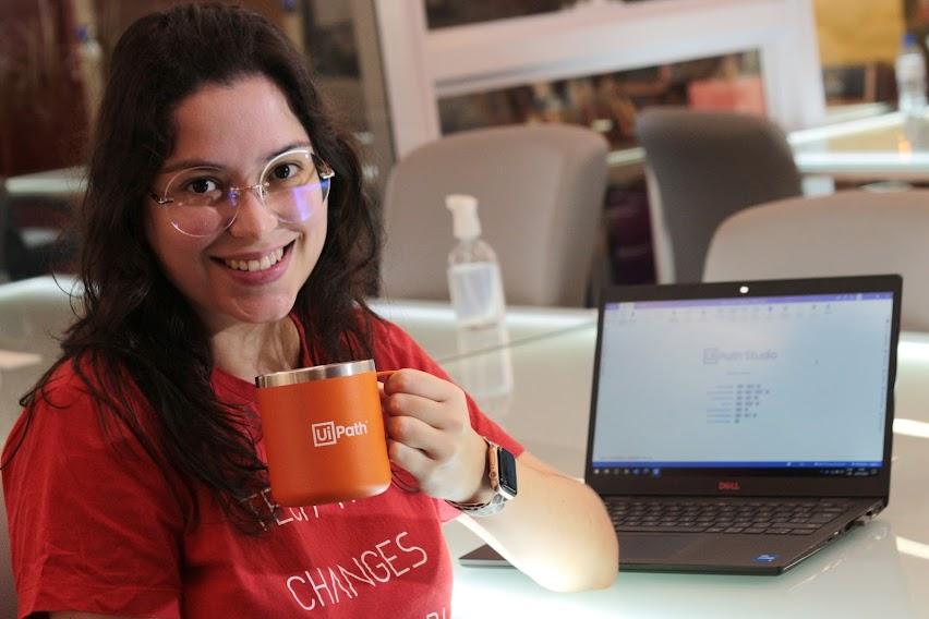 jovem sorri para a foto segurando uma caneca. Ela está sentada e há um computador pessoal atrás dela. #paratodosverem