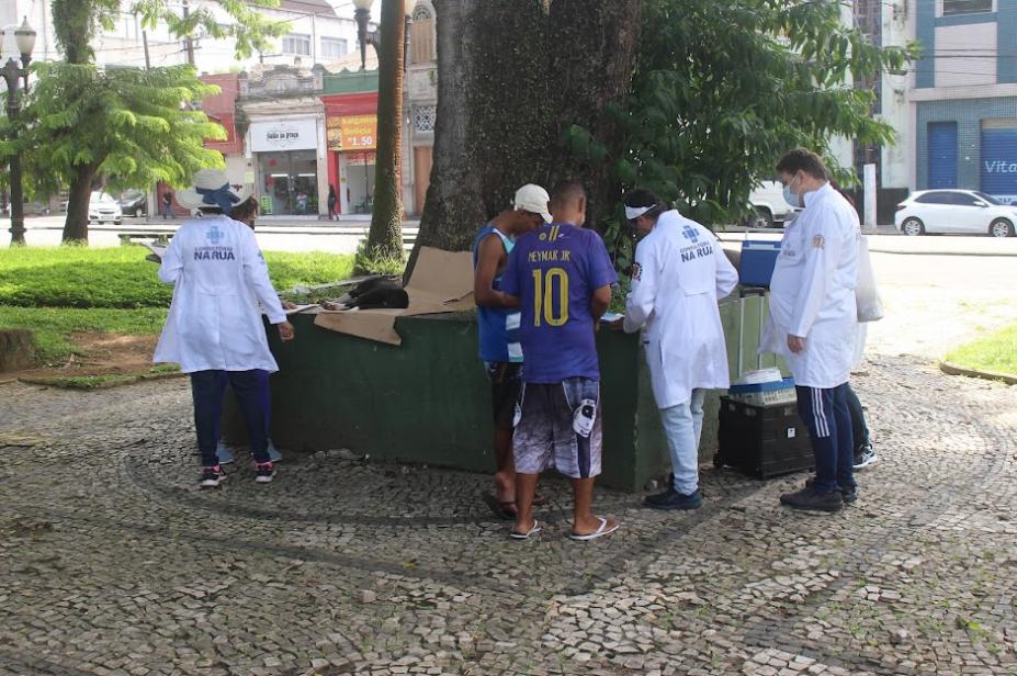 agentes usando avental branco prestam atendimento a uma pessoa em praça pública. #paratodosverem