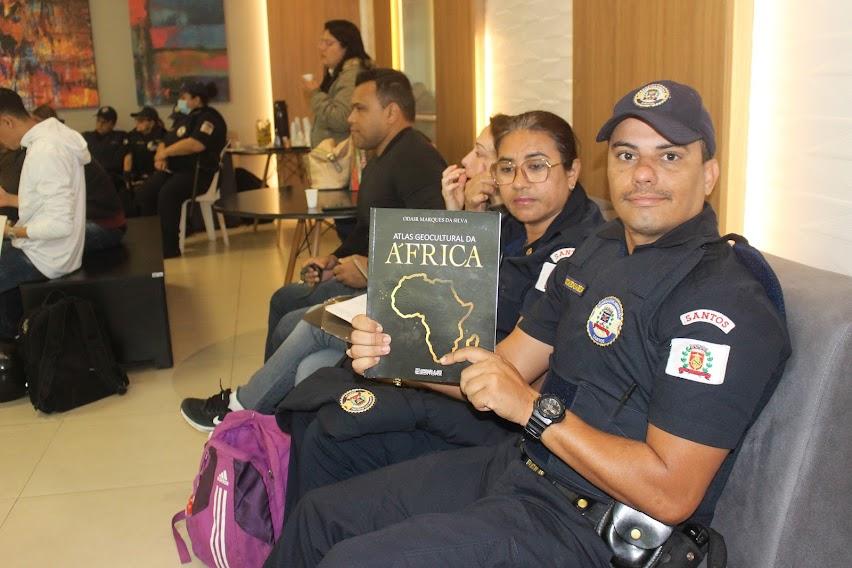 guarda municipal está sentado segurando livro em cuja capa se lê Africa e se vê o desenho do continente africano. #paratodosverem
