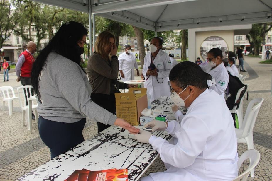 mulher faz teste de glicemia em estande com várias pessoas na praça #paratodosverem