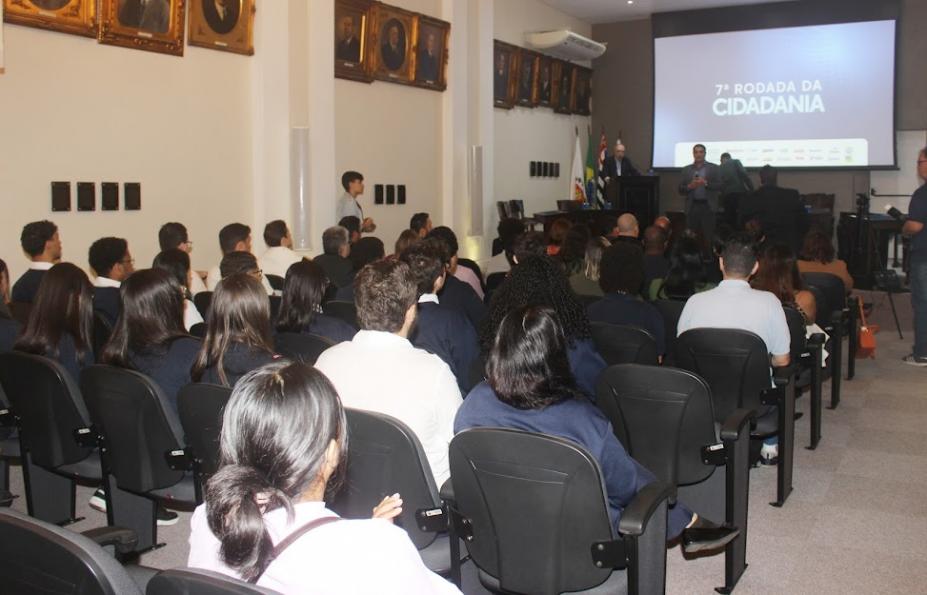 várias pessoas sentadas em auditório assistindo a uma palestra. Ao fundo há um telão onde se lê Rodada da cidadania