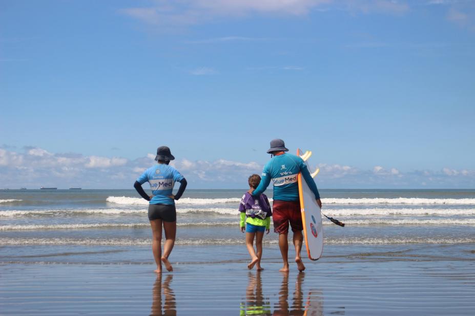 criança está entrando no mar com dois adultos. #paratodosverem
