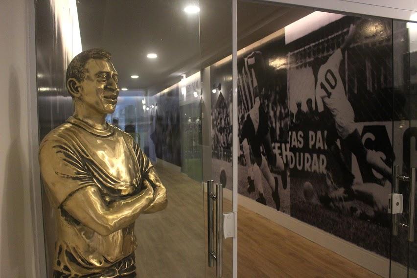 estátua de pelé está em frente a uma porta de vidro. Por dentro se vê uma parede coberta por ilustrações referentes da Pelé. #paratodosverem 