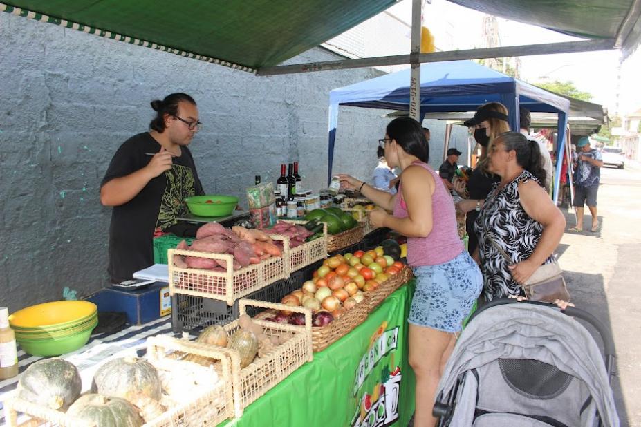 comerciante está à frente de banca de legumes. Consumidores estão olhando e tocando. #paratodosverem 