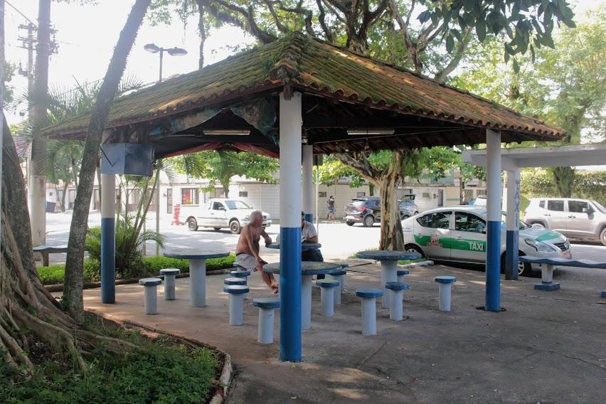 vista geral de praça com mesas e bancos. Há dois homens sentados sob o gazebo. #paratodosverem