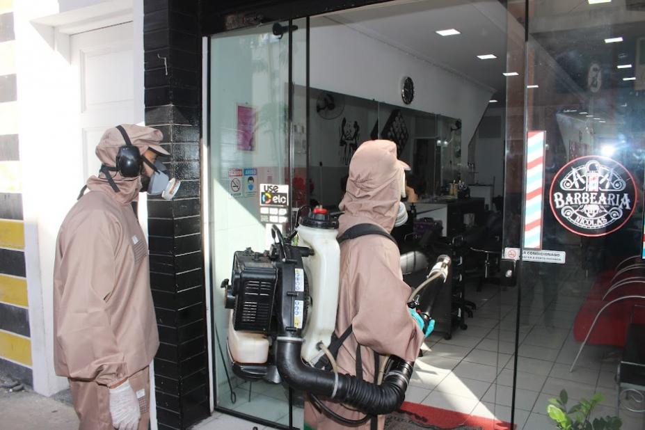 dois agentes protegidos com roupas especiais, toucas e máscara. Um deles tem um equipamento de nebulização pendurado nas costas. #paratodosverem 