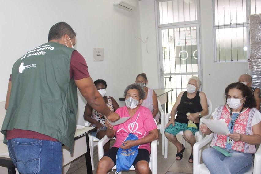 homem está entregando folheto para mulheres sentadas em sala de policlínica. #paratodosverem