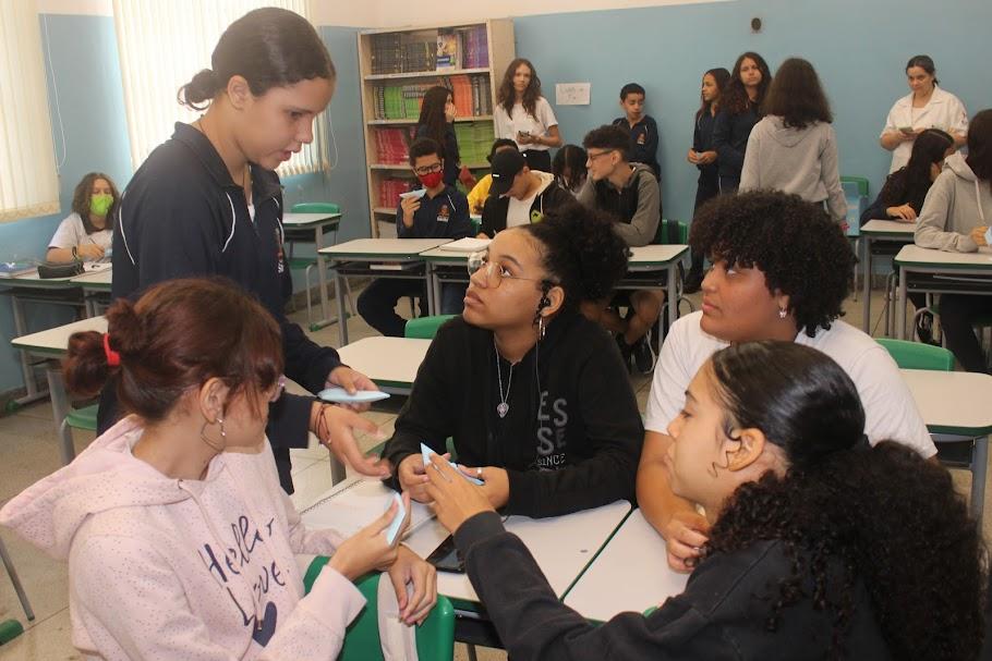 aluna em pé distribui papeis para meninas sentadas em sala de aula. #paratodosverem