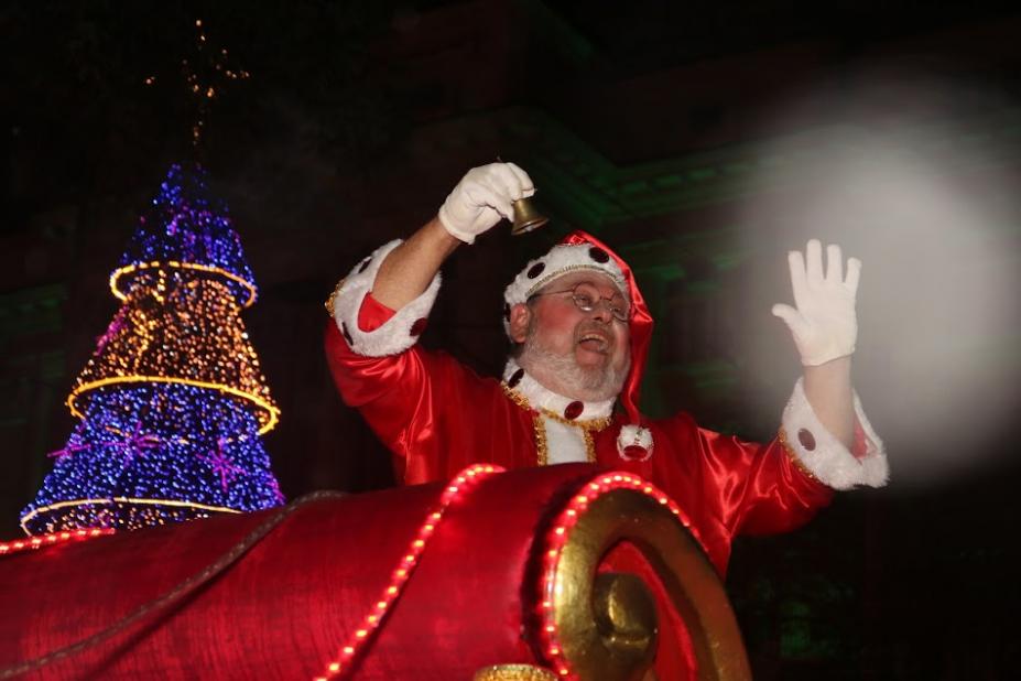 papai noel passa de treno diante de grande árvore de natal iluminada. Ele toca o sino e saúda o público (que não aparece na imagem). Ao fundo, o paço municipal. #paratodosverem