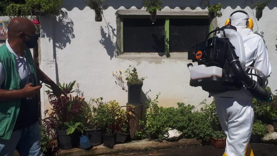 dois homens estão na frente de uma casa. Um deles faz nebulização com um equipamento acoplado às costas. O outro acompanha. #paratodosverem