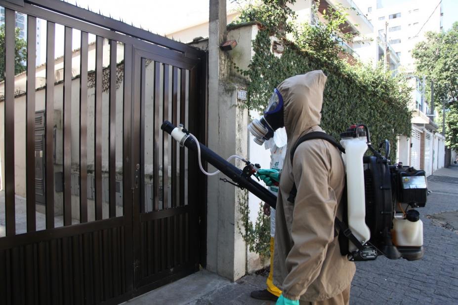 Homem paramentado com macacão, máscara e equipamento nebulizador nas costas. #Paratodosverem