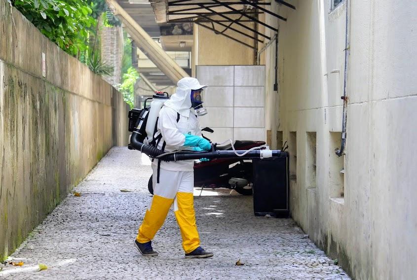 homem paramentado com calça, capa, capacete coberto e luvas, tem pulverizador nas costas. #paratodosverem