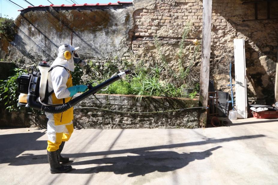 Homem com equipamento acoplado às costas pulveriza ambiente aberto. #Paratodosverem