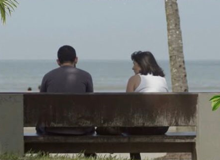 casal está sentado em banco da praia, de frente para o mar e de costas para a foto. #paratodosverem 