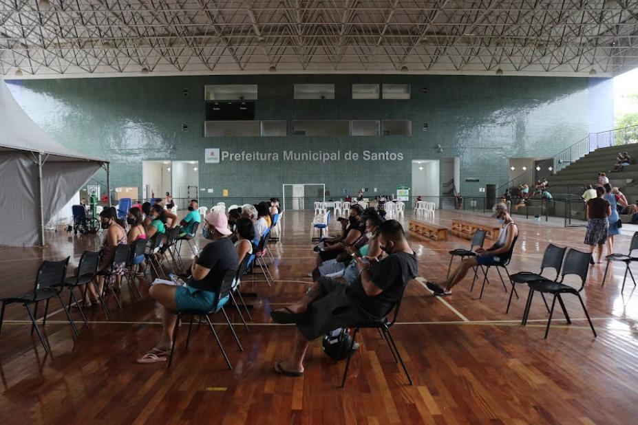 ginásio coberto com cadeira distanciadas e pessoas sentadas. Ao fundo se lê prefeitura municipal de santos. #paratodosverem