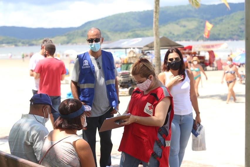 Voluntária da Cruz Vermelha, em pé, conversa com casal sentado em banco e anota dados em prancheta. #pracegover