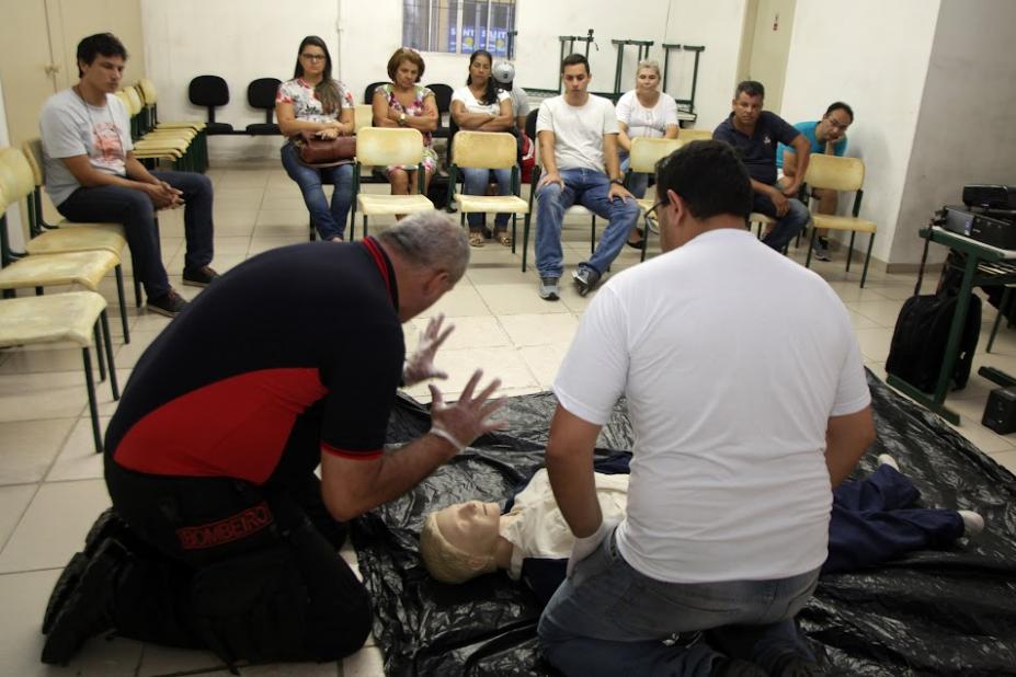 dois homens ajoelhados no chão simulam atendimento a  pessoa ferida. Trata-se de um boneco deitado também no chão. Ao fundo, os alunos atentos. #paratodosverem