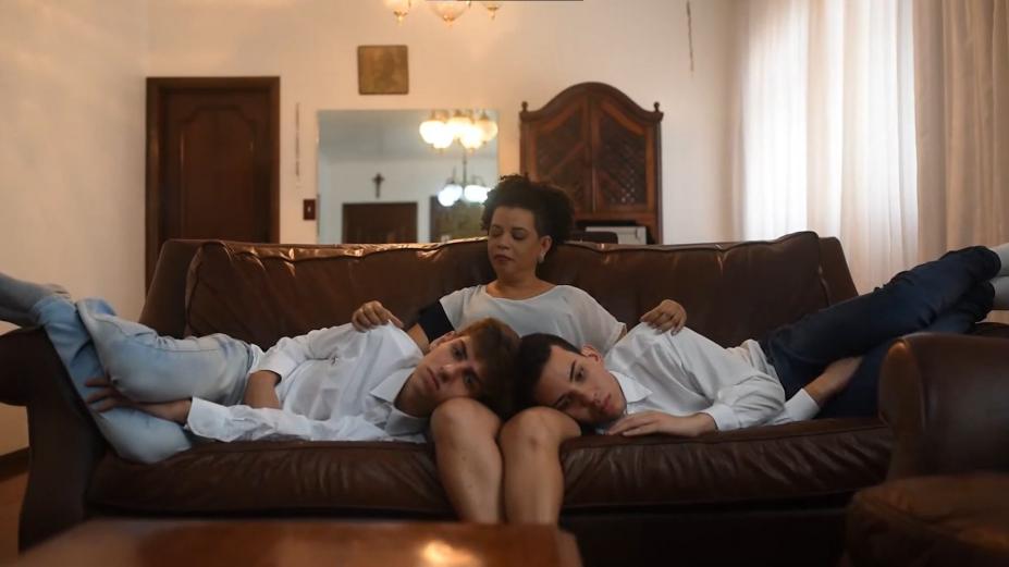 Dois jovens estão deitados em um sofá, de lados opostos, ambos com a cabeça no colo de uma mulher. #Paratodosverem