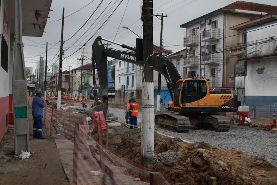 rua com obras. Retroescavadeira em movimento e homens trabalhando. há terra nos canteiros e a pista está coberta  por pedras. #paratodosverem