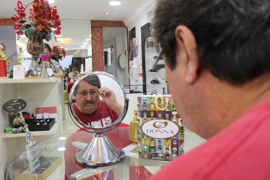 homem está de costas para foto se olhando num espelho e segurando a armação do óculos no rosto. #paratodosverem