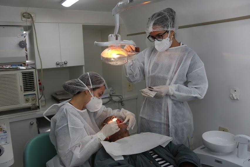duas dentistas atendem homem em cadeira própria para serviço dentário. Ambiente é um consultório.  #paratodosverem