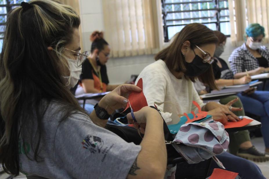 educadoras recortam figuras e desenham em papel. Elas estão sentadas em uma sala de aula. #paratodosverem