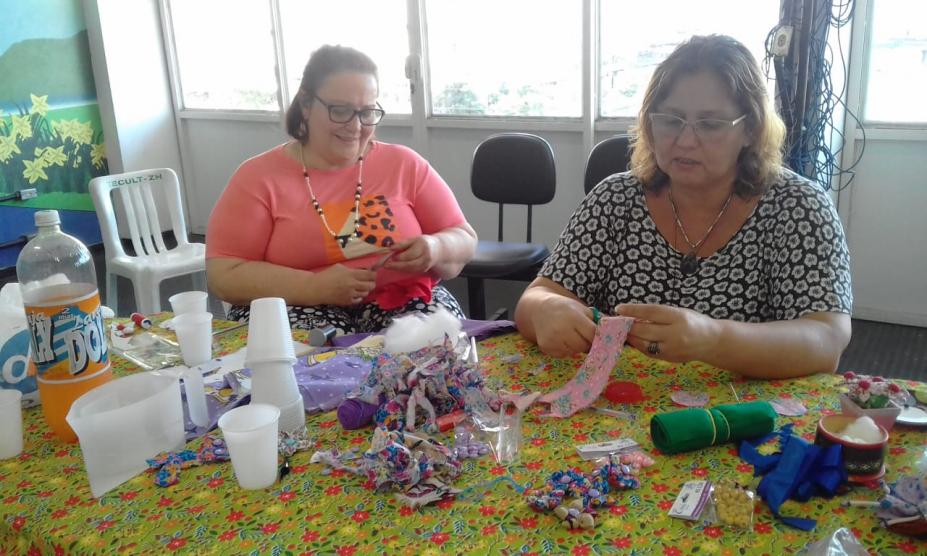 Mulheres estão sentadas e recortando e costurando. Sobre a mesa à frente delas há produtos para artesanato, como tecidos, fitas, linha e tesoura. #Pracegover