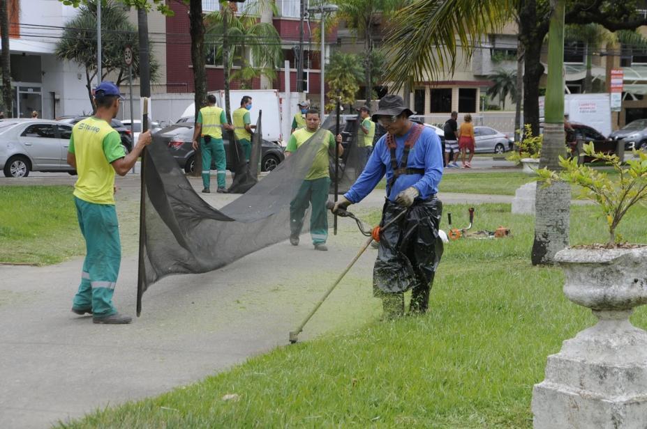 homens estão cortando grama no jardim da praia. Em primeiro plano, um deles usa uma máquina para cortar a grama. Ao fundo, dois seguram uma rede de proteção para a grama cortada não espalhar. #paratodosverem