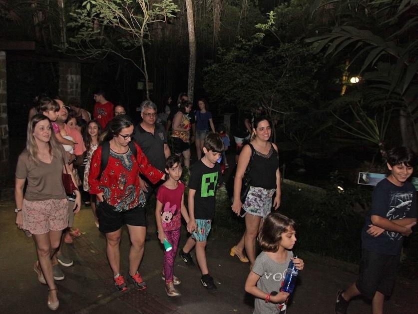 vários adultos e crianças caminhando por alameda do orquidário à noite. #paratodosverem